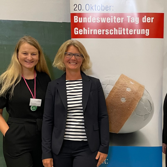 Dr. Susanne Schaefer und Dr. med. Michaela Veronika Bonfert posieren vor einem Banner zum Tag der Gehirnerschütterung